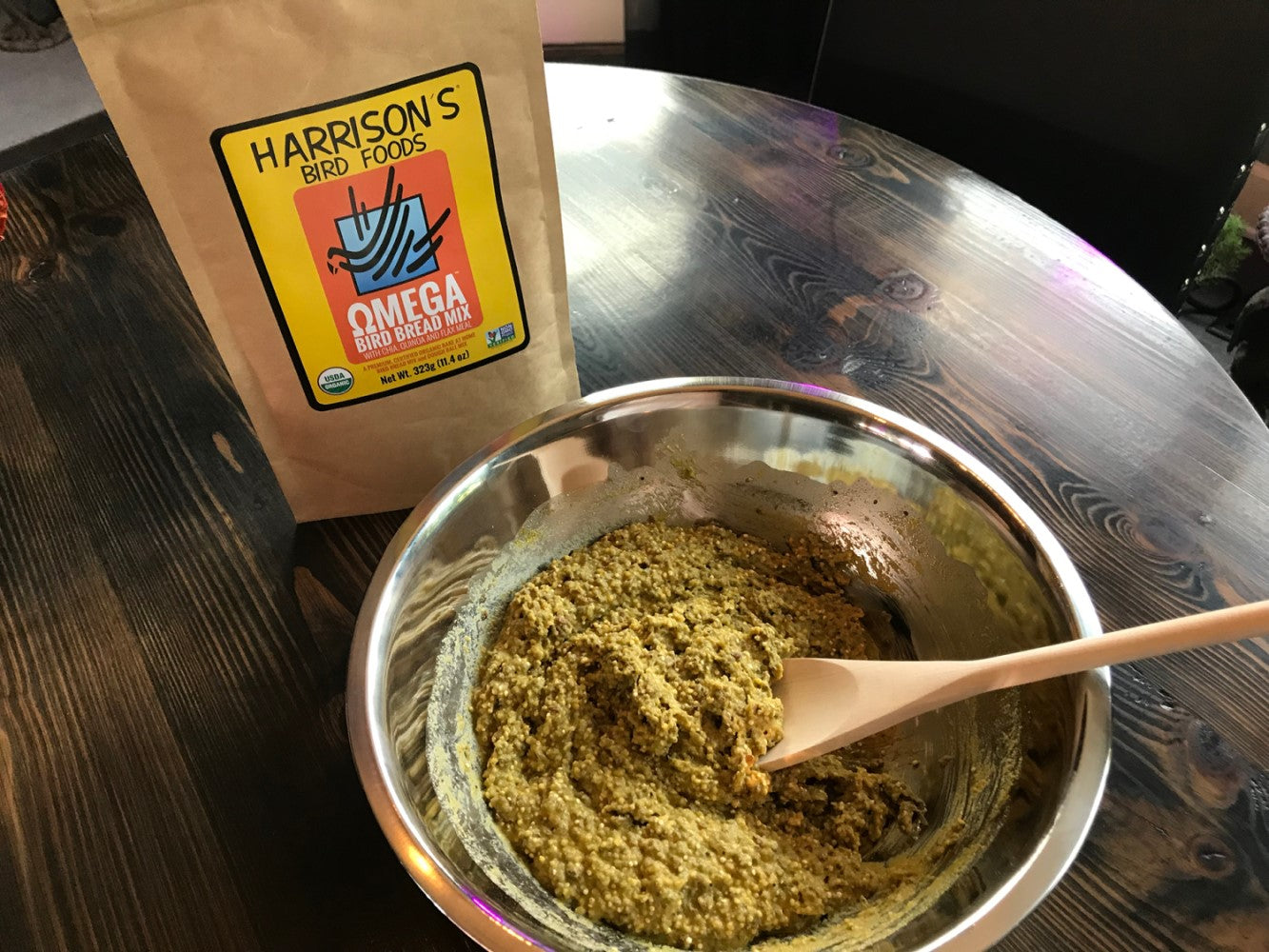 A bag of Omega Harrison's Bird Bread next to a bowl in which the mix is being stirred with a wooden spoon